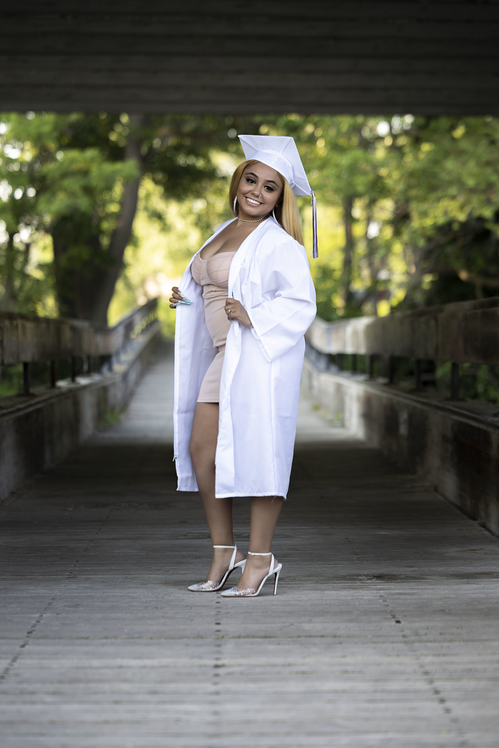 Senior girl in white cap and gown in a nude colored dress and high heels in the summer on a bridge