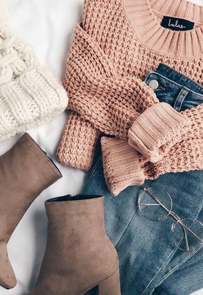 An outfit laid out on a bed with brown booties, wire frame round glasses, jeans, blush pinks sweater, and white knit beanie hat