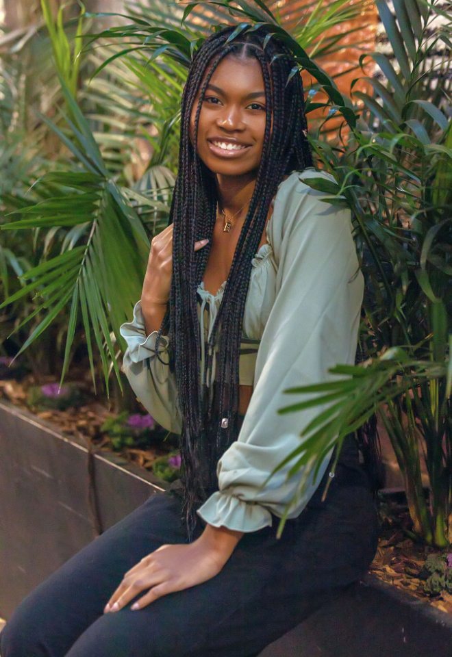 Senior girl with braids wearing sea green long sleeve crop top and black destructed jeans sitting on ledge with plants in the background