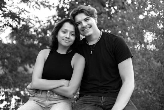 Senior girl in a black tank top and blue jeans and senior boy in a black t-shirt and jeans sitting on a concrete half wall