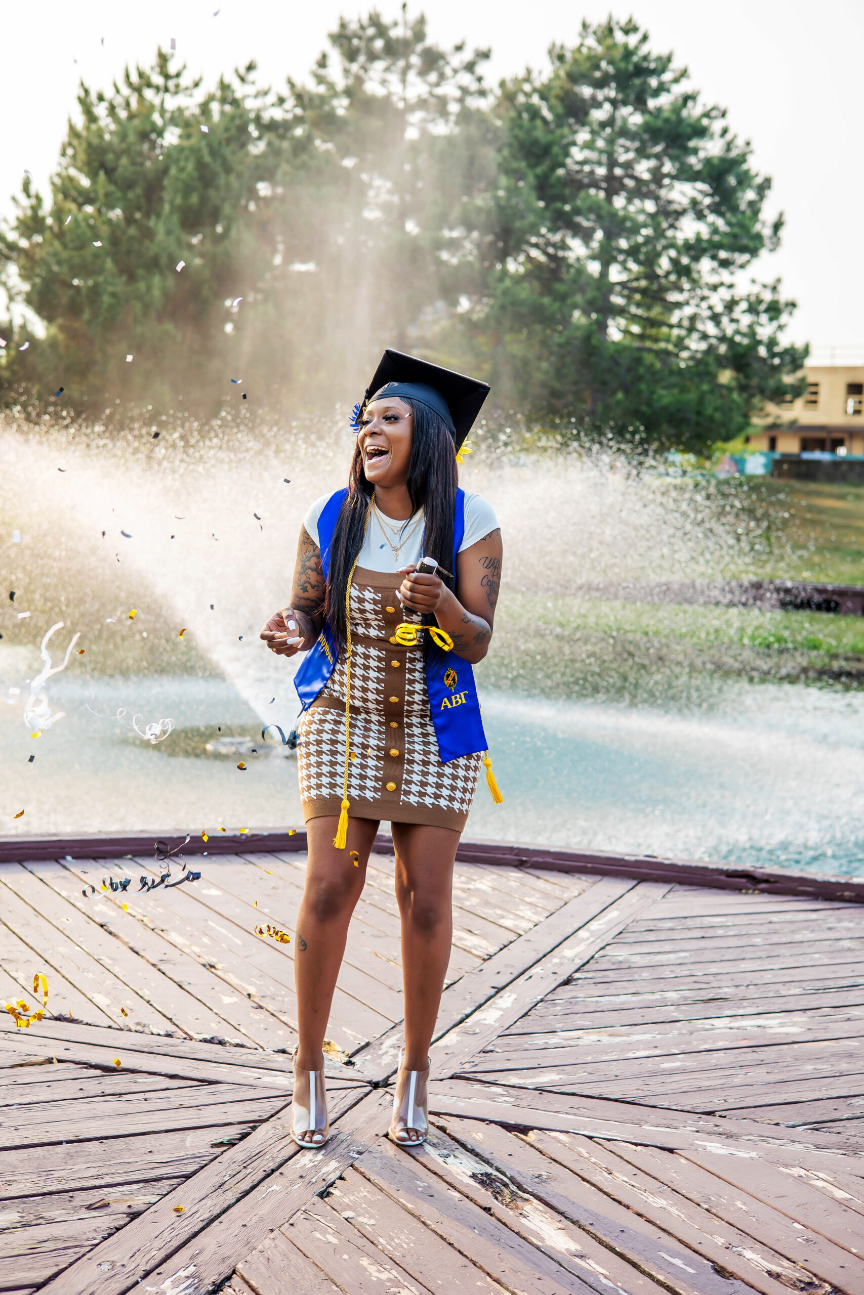 College grad wearing black graduation cap, blue honors sash, yellow cord and brown and white dress posing in front of a pond. Ypsilanti senior & college grad photographer