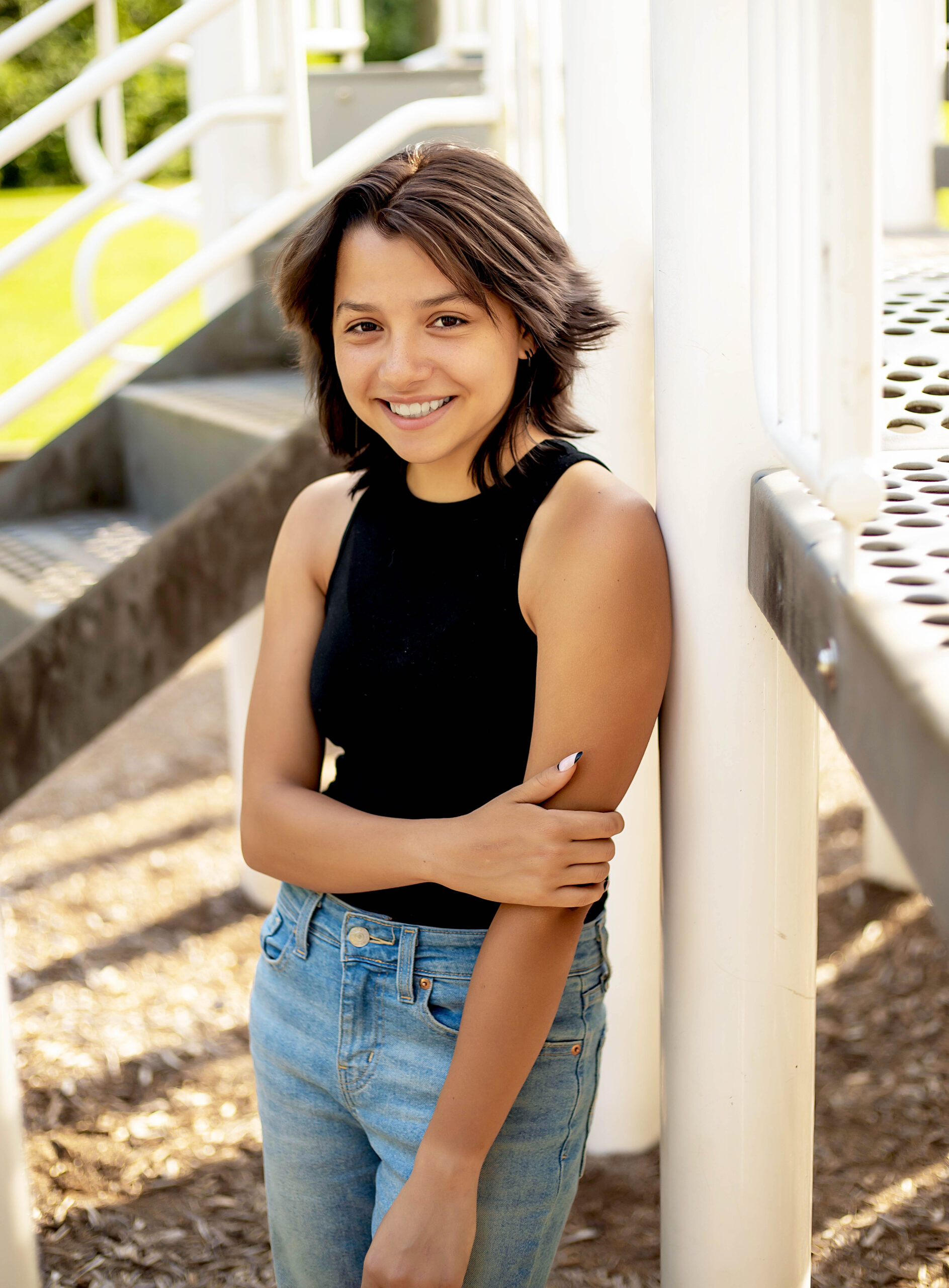 Senior girl in black tank top and blue jeans leaning against a pole in a park